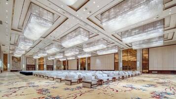 A spacious conference room with rows of white chairs, a patterned carpet, and a high ceiling with chandeliers.