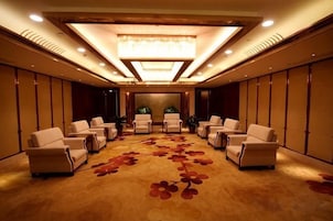 Un salón de conferencias con piso alfombrado, múltiples sillas dispuestas en filas, una araña de cristal y una planta en el centro.