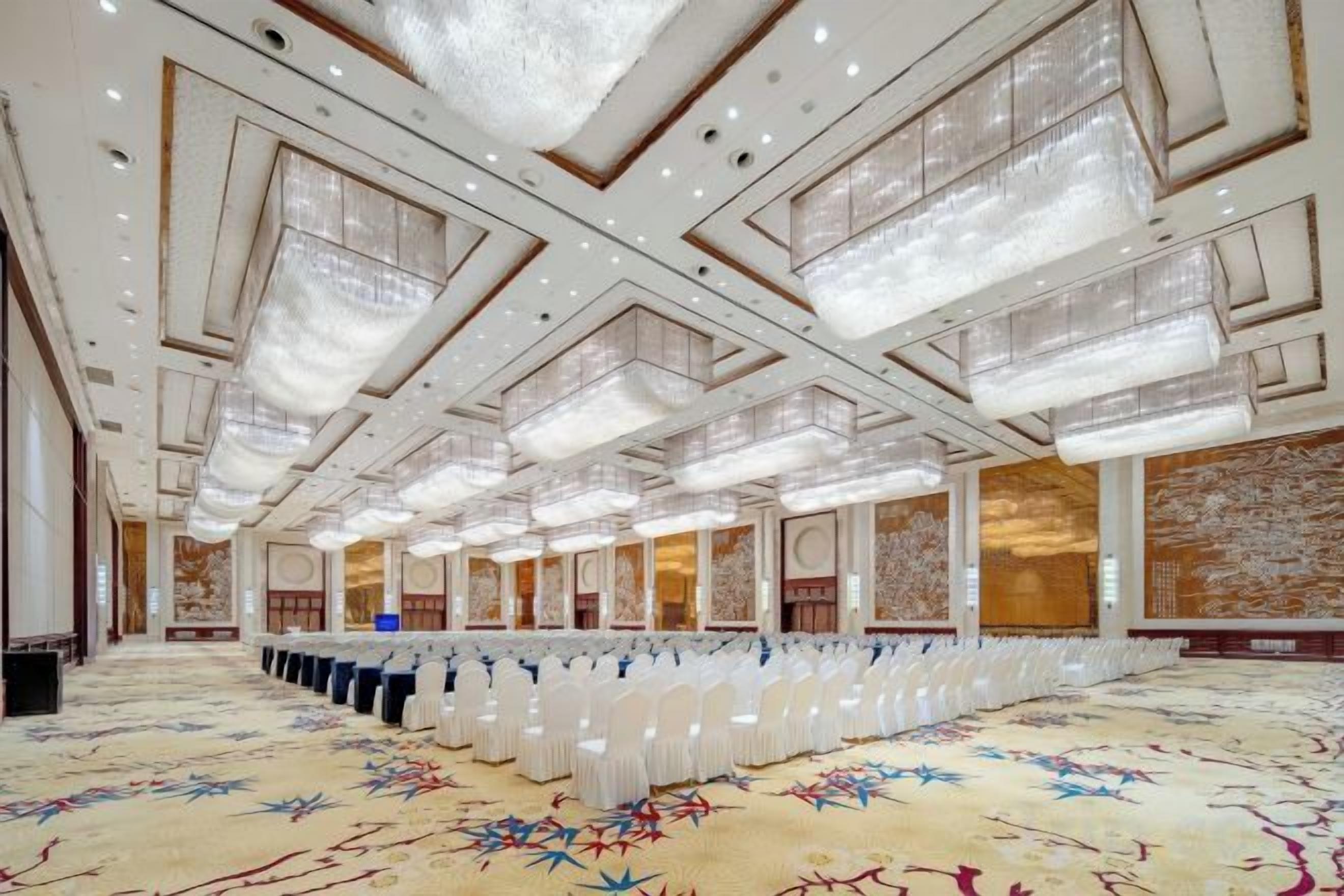 A large conference room with rows of white chairs, a patterned carpet, and a high ceiling with chandeliers.