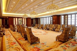 Un salón de conferencias con filas de sillas, una alfombra grande con motivos florales y candelabros.