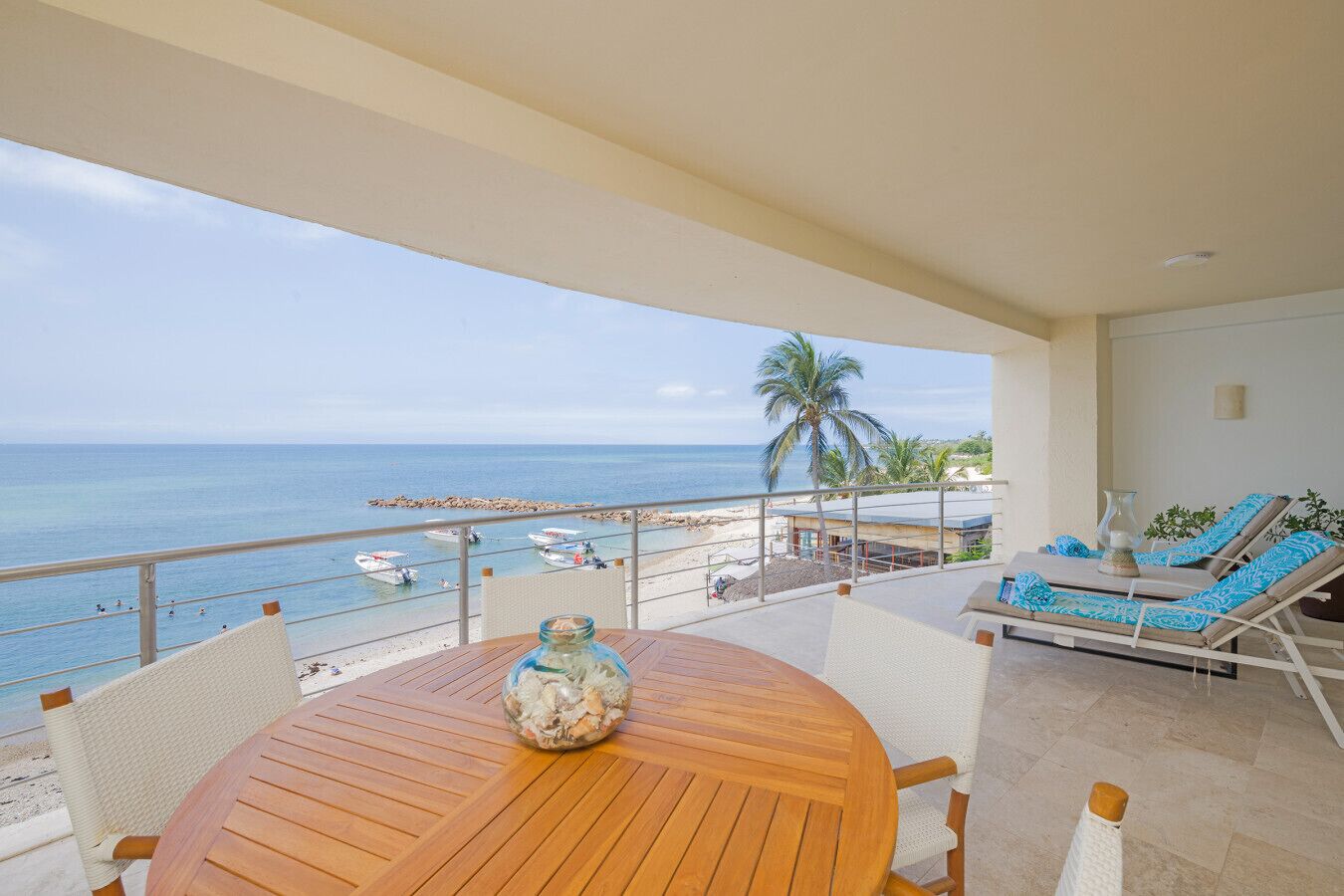 Un balcón con mesa y sillas de madera, con vista a una playa con barcos y palmeras.