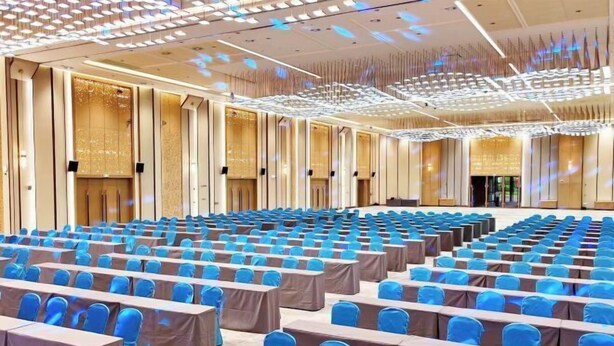 Un amplio salón de conferencias con filas de mesas y sillas, un diseño moderno de techo y una gran pantalla al frente.