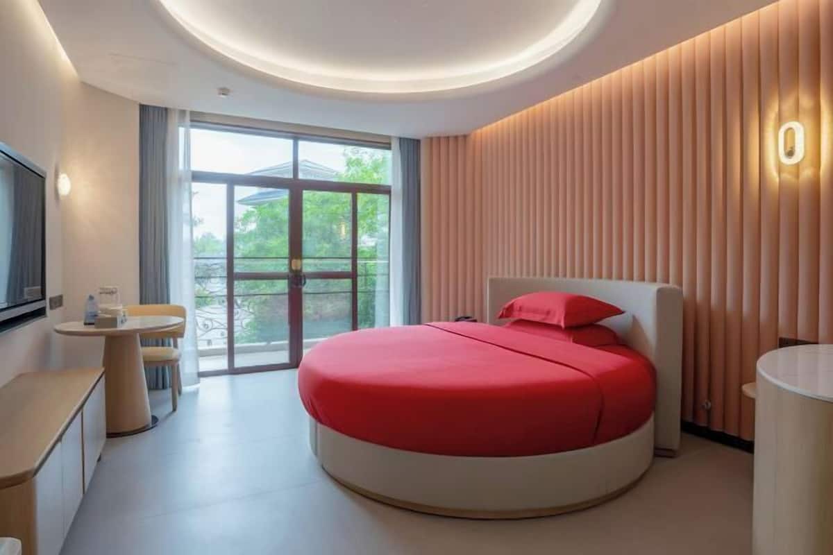 Habitación de hotel moderna con una cama redonda, una pequeña mesa circular, un televisor montado en la pared y un balcón con vistas.