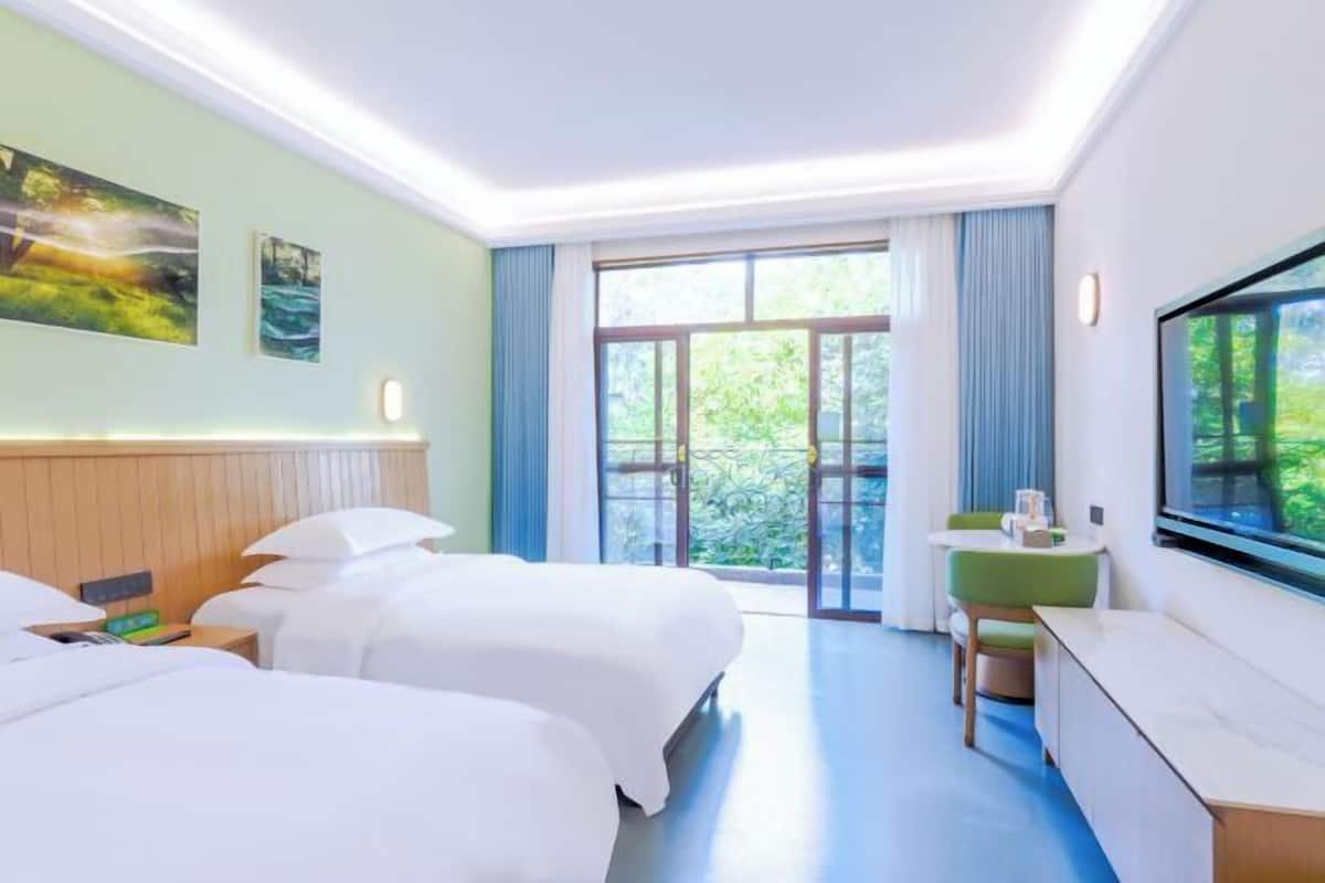 Habitación de hotel con dos camas, un escritorio, un televisor y un ventanal con vistas a zonas verdes.