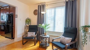 A living room with two chairs, a small table with a plant, a window with curtains, and a kitchen area with a refrigerator. - Spacious - 3 bedrooms house in Granby, Quebec (Granby)