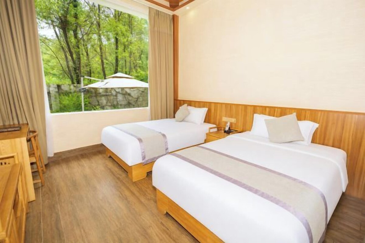 A hotel room with two beds, a desk, and a window with a view of trees.