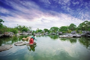 A serene outdoor setting with a person in traditional attire standing on rocks in a body of water, surrounded by greenery and rocks.