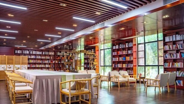 A modern library with wooden bookshelves, a long table with chairs, and large windows allowing natural light. - Stone Hotel (Yingde)