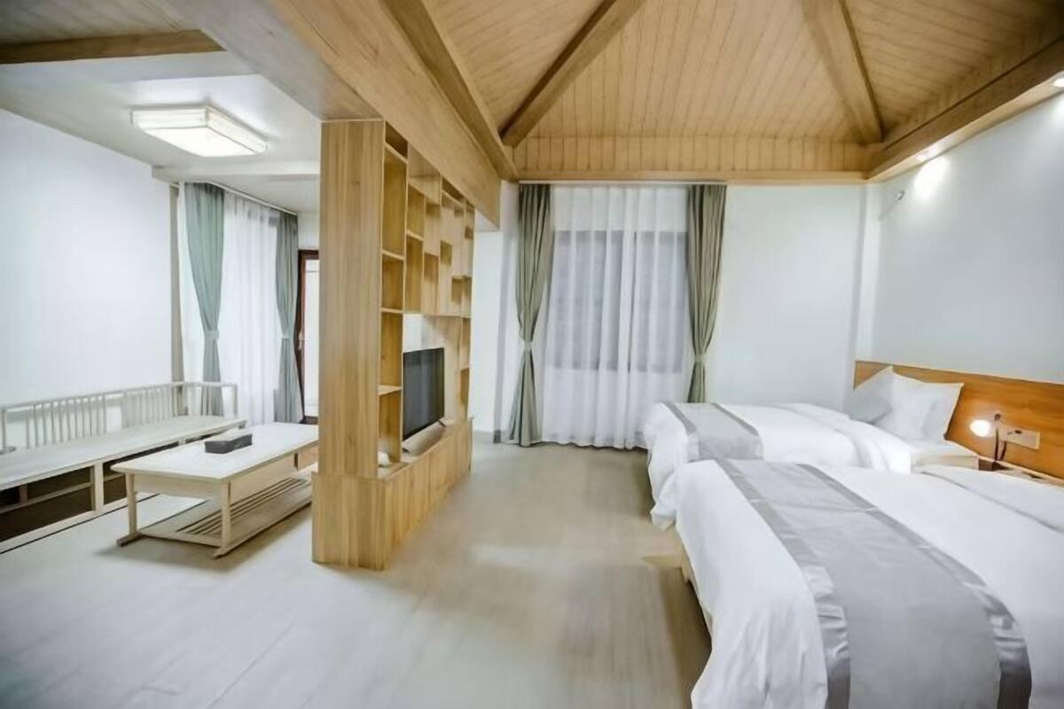 A hotel room with a wooden ceiling, a TV on a wooden stand, two beds, a desk, and a window with curtains.