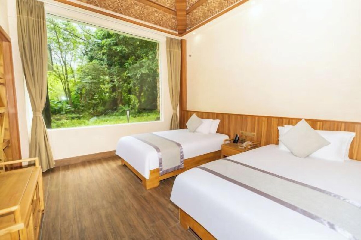 A hotel room with two beds, a large window with a view of greenery, and a wooden ceiling.