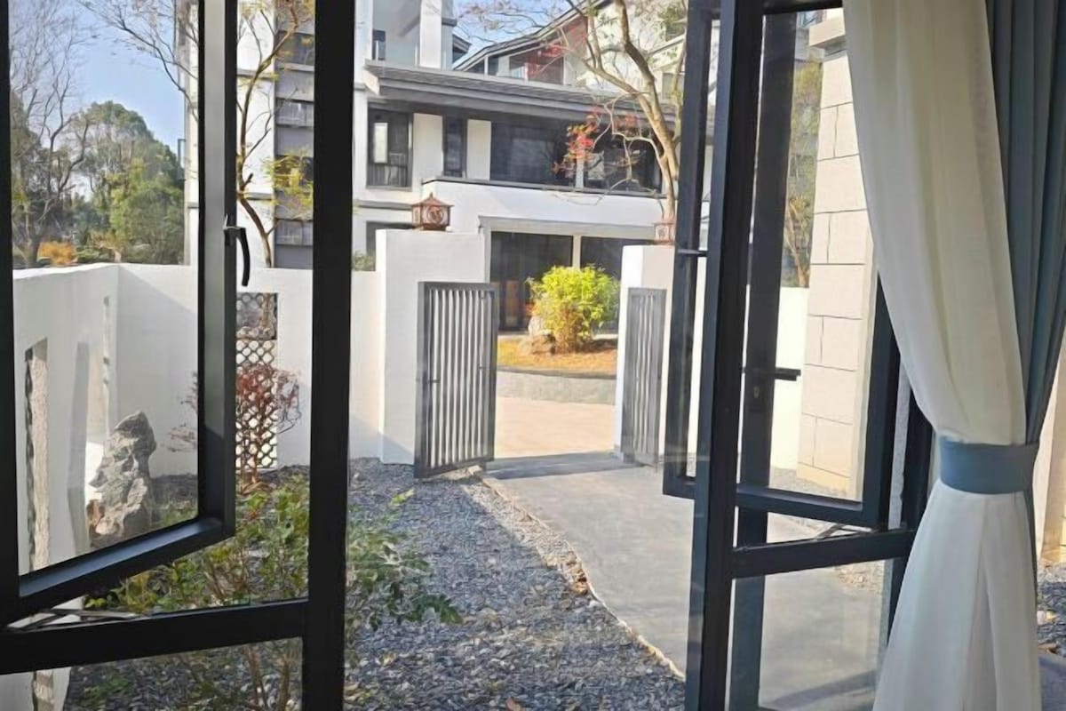 A modern building with a gated entrance, a small garden area, and a view of a residential building in the background.