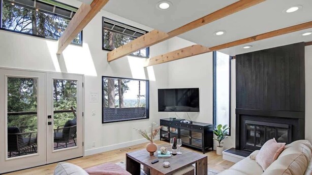 A modern living room with a fireplace, a flat-screen TV, and a large window overlooking a deck. - Forest-View Hot Tub • 5 Min to UO • Game Room (Eugene)