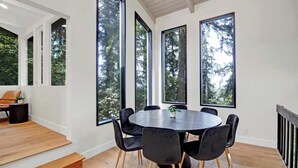 A modern dining area with a round table and black chairs, large windows with a forest view, and a wooden floor. - Forest-View Hot Tub • 5 Min to UO • Game Room (Eugene)