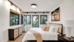 A modern bedroom with a large bed, a bedside table, a nightstand, a dresser, and a window with a view of trees. - Forest-View Hot Tub • 5 Min to UO • Game Room (Eugene)