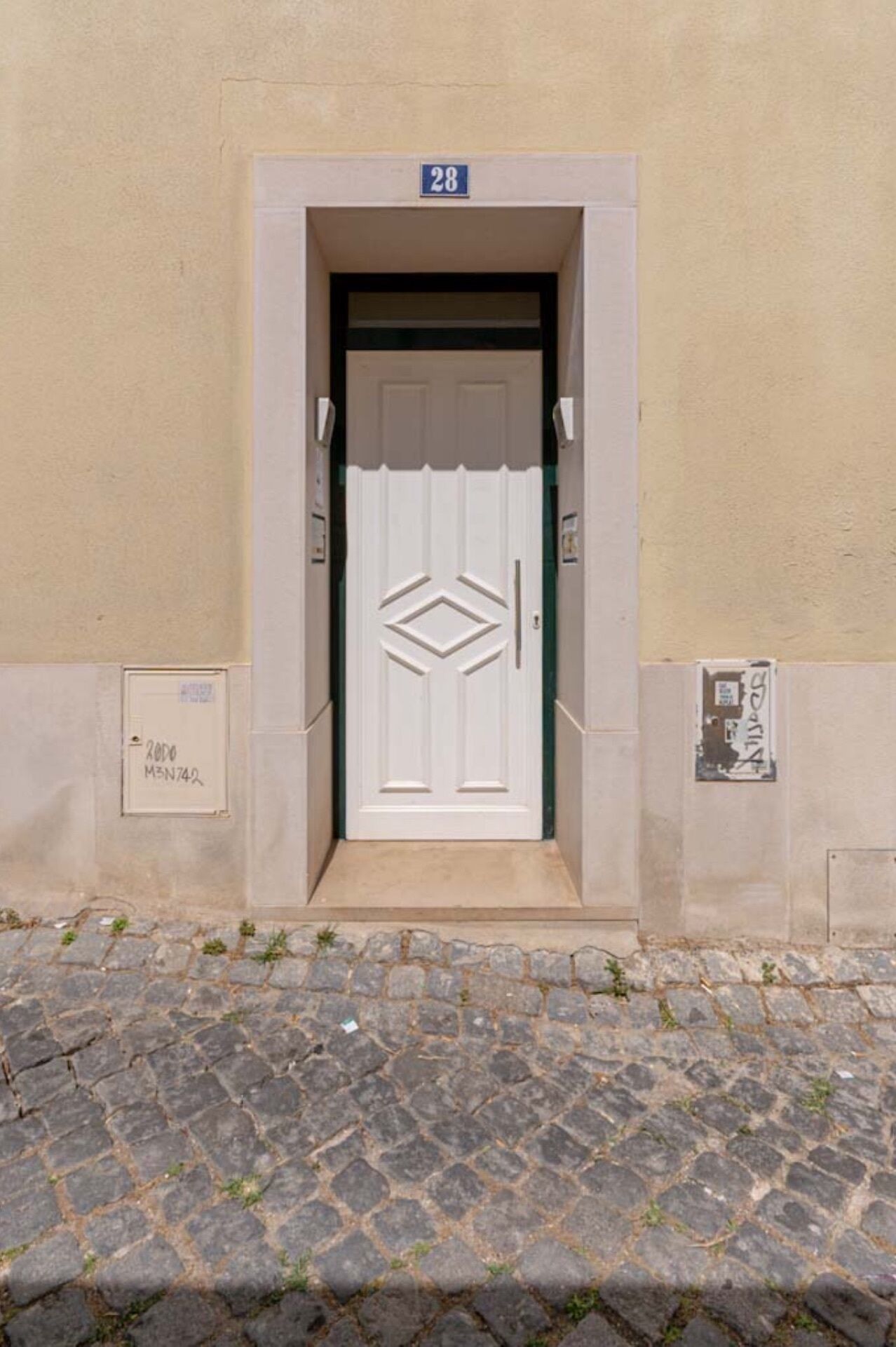 A building entrance with a white door, a number '28' above it, and a cobblestone pavement in front.