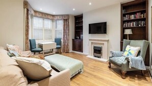 A living room with a sofa, armchair, TV, fireplace, bookshelf, and a desk with chair.
