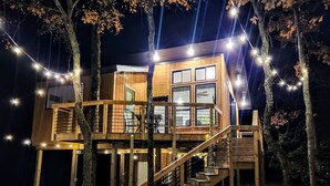 Uma cabana de madeira com escada que leva a um deck, cercada por árvores e luzes de Natal.