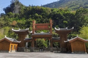 A traditional gate with a sign, flanked by two wooden buildings, set against a backdrop of a forested hill.