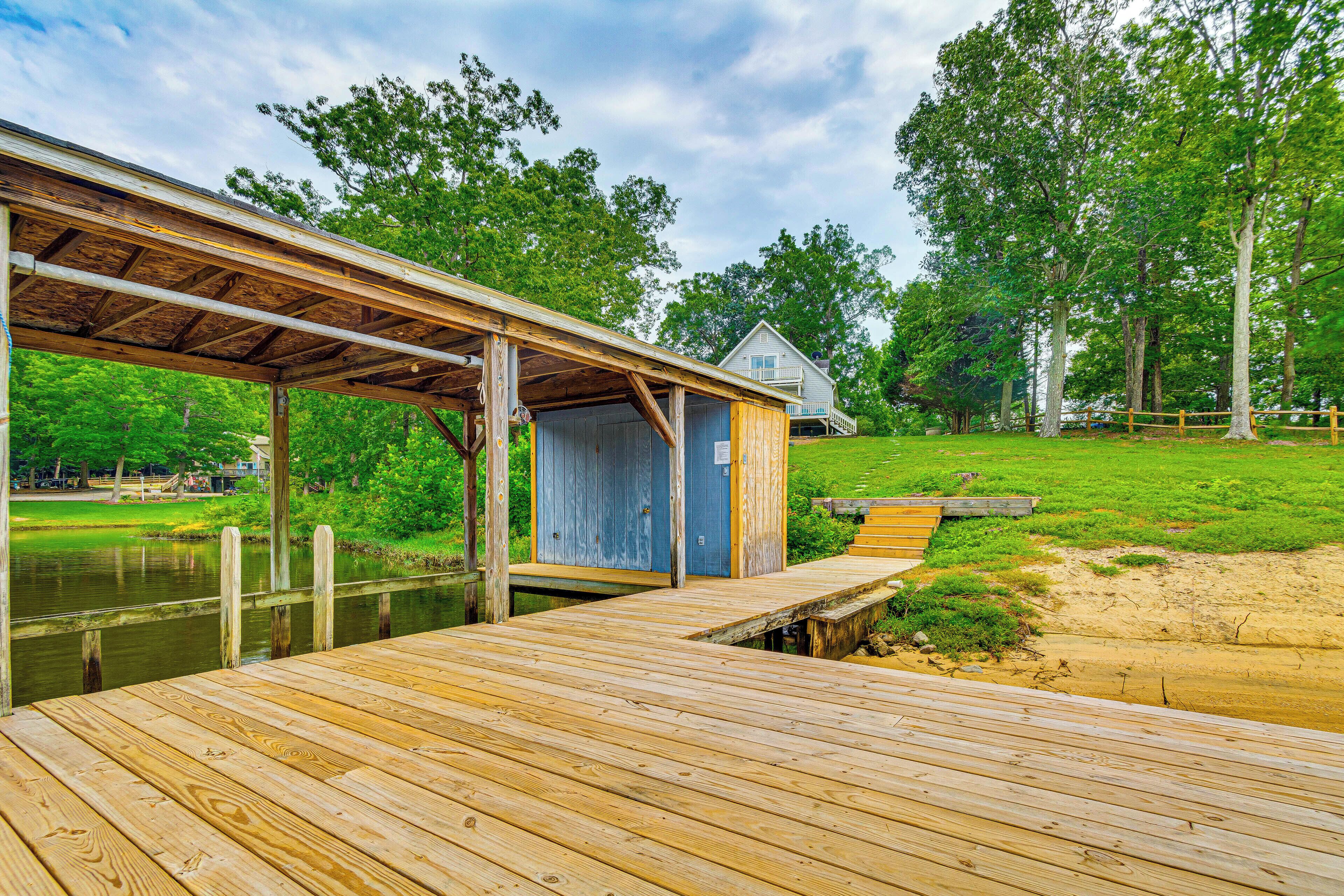 A wooden dock leading to a covered structure with a blue door, a grassy area, and a house in the background.