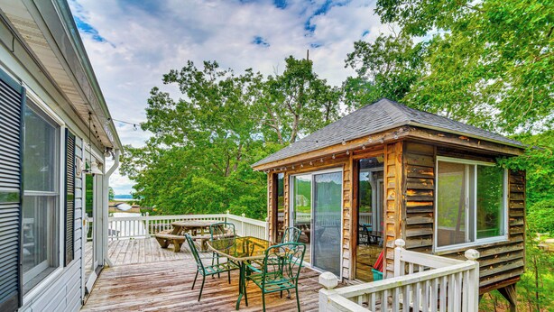 A wooden deck with a glass enclosure, outdoor furniture, and a view of a forested area. - Private Island Getaway in Roanoke Rapids w/ Grill! (Roanoke Rapids)