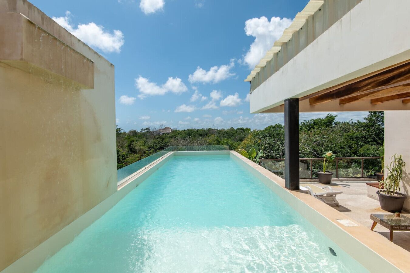 A rectangular swimming pool with clear water, surrounded by a concrete deck and a covered area with seating.
