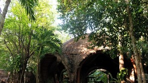 Une construction traditionnelle à toit de chaume, dotée de plusieurs arcades et entourée d’une végétation luxuriante et d’arbres.