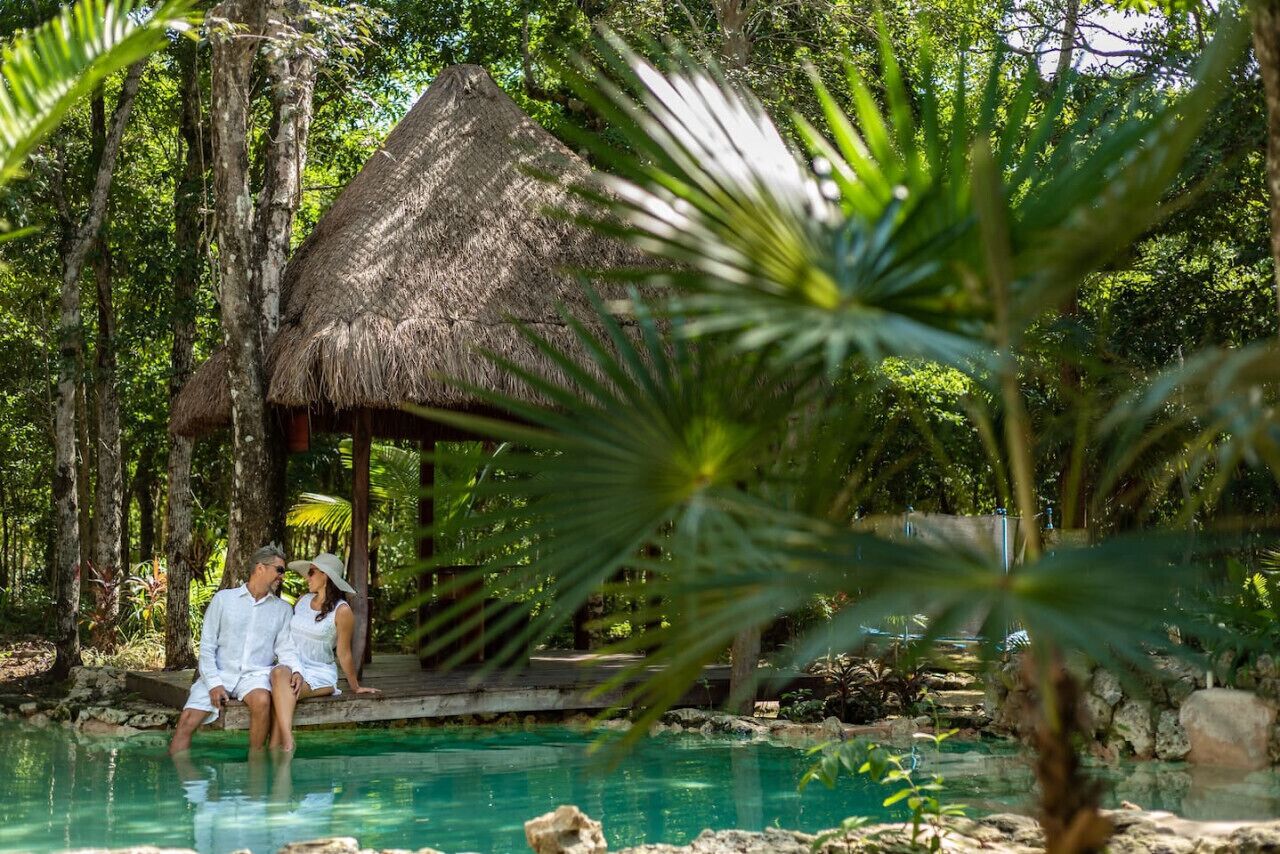 A couple sitting by a pool with a thatched-roof hut in a lush, tropical setting.
