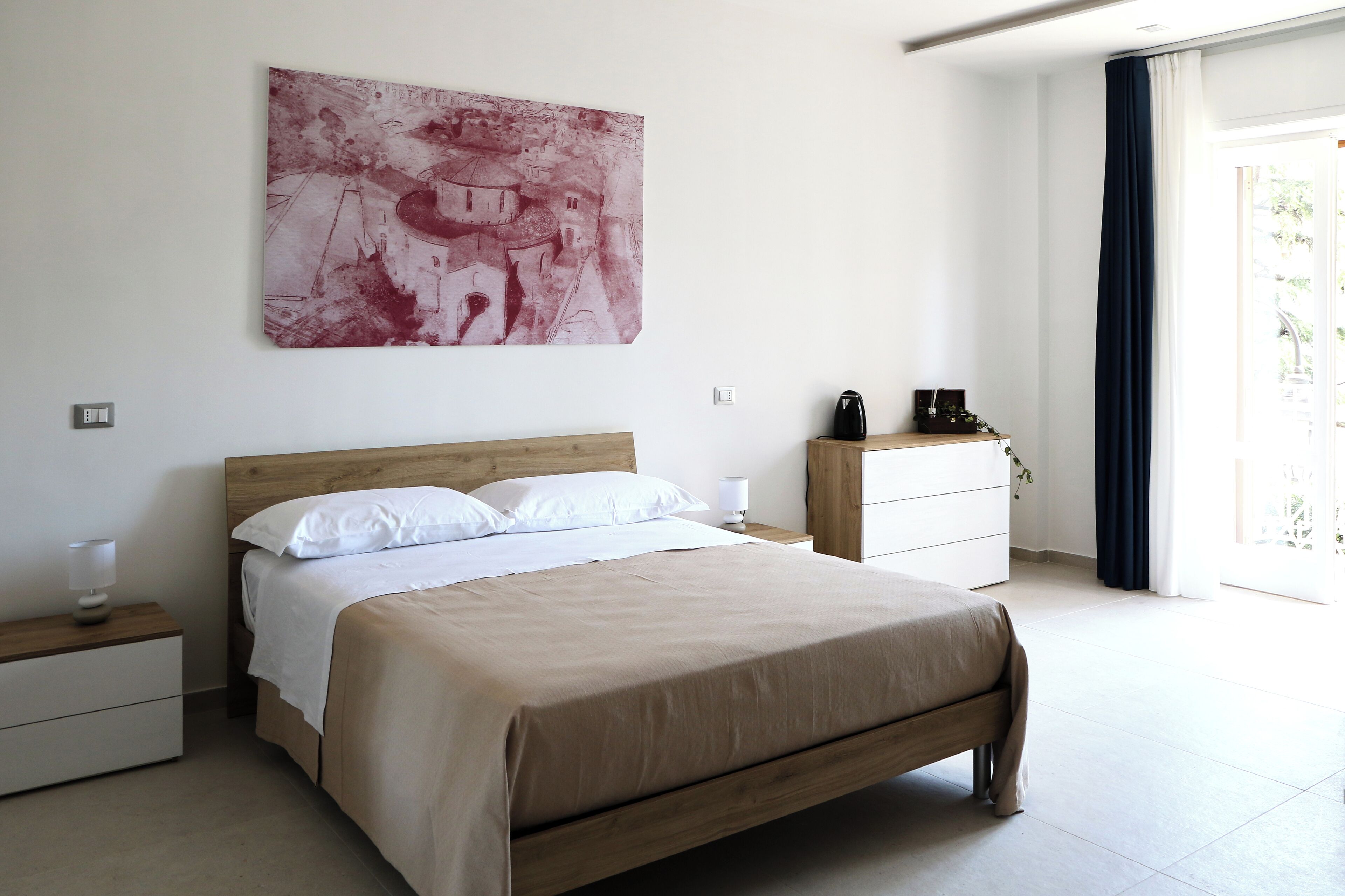 A modern bedroom with a wooden bed, white bedding, a nightstand with a lamp, a dresser, a coffee maker, and a large abstract painting on the wall.