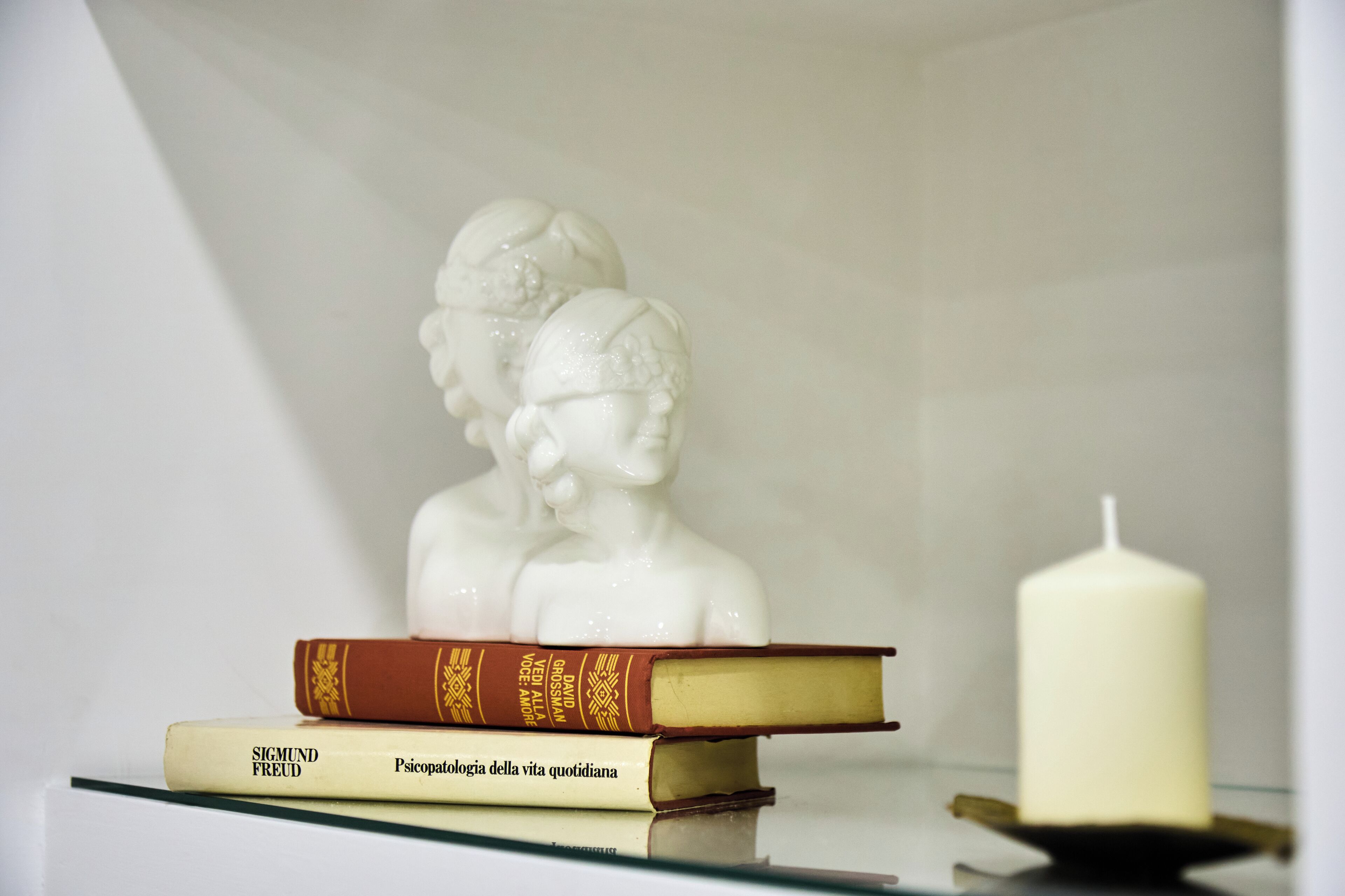 A white bust sculpture, a stack of books, and a white candle on a glass shelf.