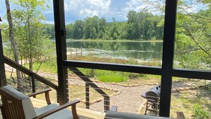 Een overdekt terras met houten stoelen en een rustieke houten kruk, met uitzicht op een meer en een bebost landschap.
