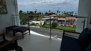 A balcony with a view of the sea and palm trees, featuring a ceiling fan, a table with chairs, and a painting on the wall. - Luxury Condo at Viva Jaco Residence (Jaco)