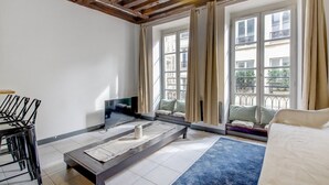 A room with a wooden ceiling, a large window with curtains, a sofa, a TV, and a small table. - Paris Marais Lombards (Paris)