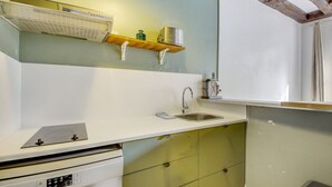 A kitchen with a white countertop, stainless steel sink, and a built-in oven. There are wooden shelves above the countertop and a window with curtains. - Paris Marais Lombards (Paris)
