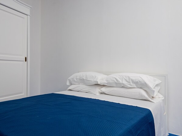 A bed with white bedding and a blue blanket, a white door, and a plain wall. - De Vivo Realty - Camilla House Positano (Positano)
