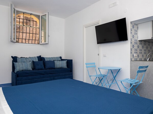A room with a bed, a sofa, a TV mounted on the wall, a small table with chairs, and a window with a view of a building. - De Vivo Realty - Camilla House Positano (Positano)