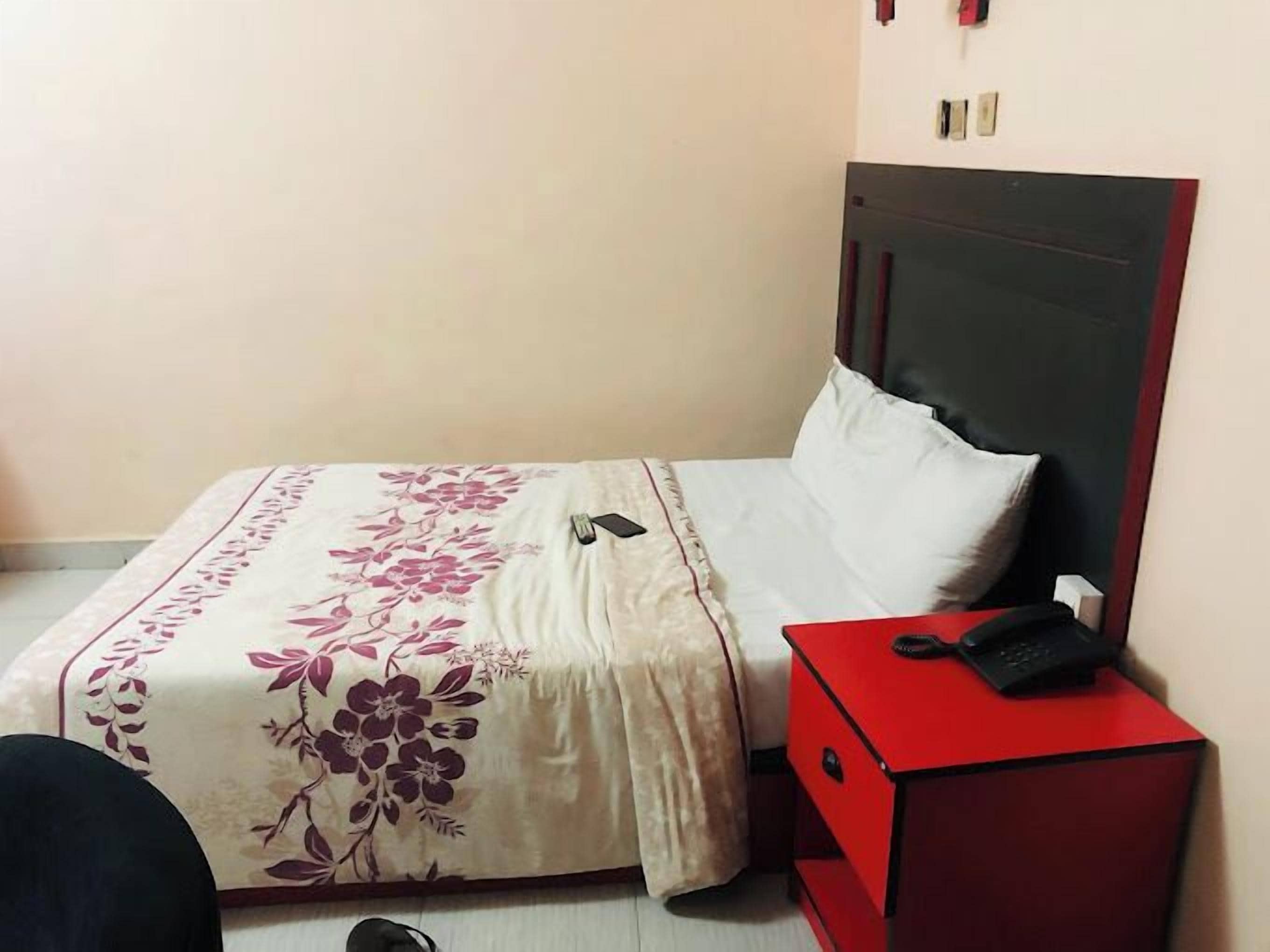 Una cama con cubrecama floral, almohada blanca y mesita de noche roja con un teléfono.