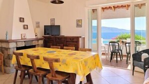 Un coin repas avec une table recouverte d’une nappe jaune, des chaises en bois, une cheminée en pierre et un téléviseur posé sur un meuble en bois.