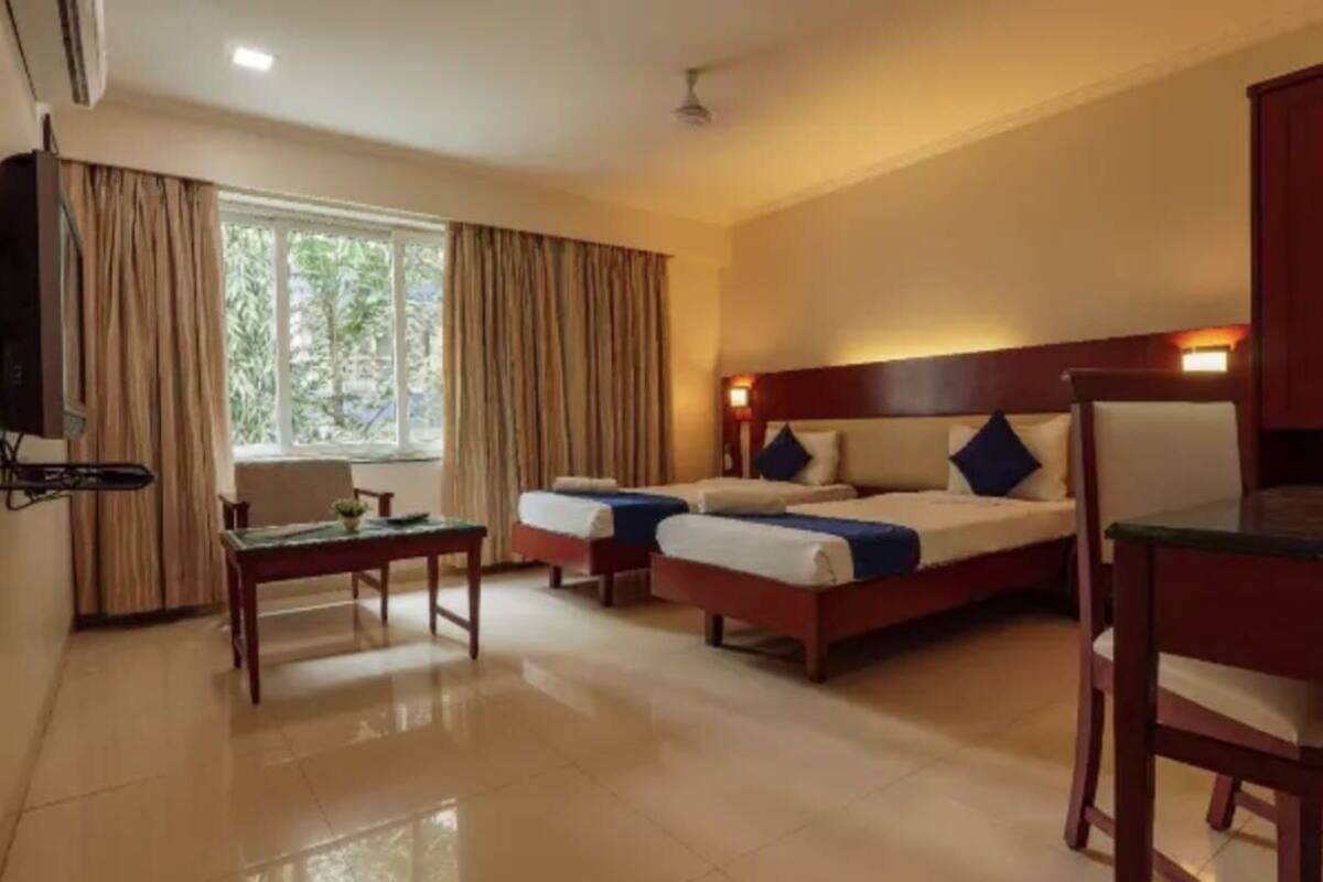 A hotel room with two single beds, a small table, a chair, a TV, and a window with curtains.