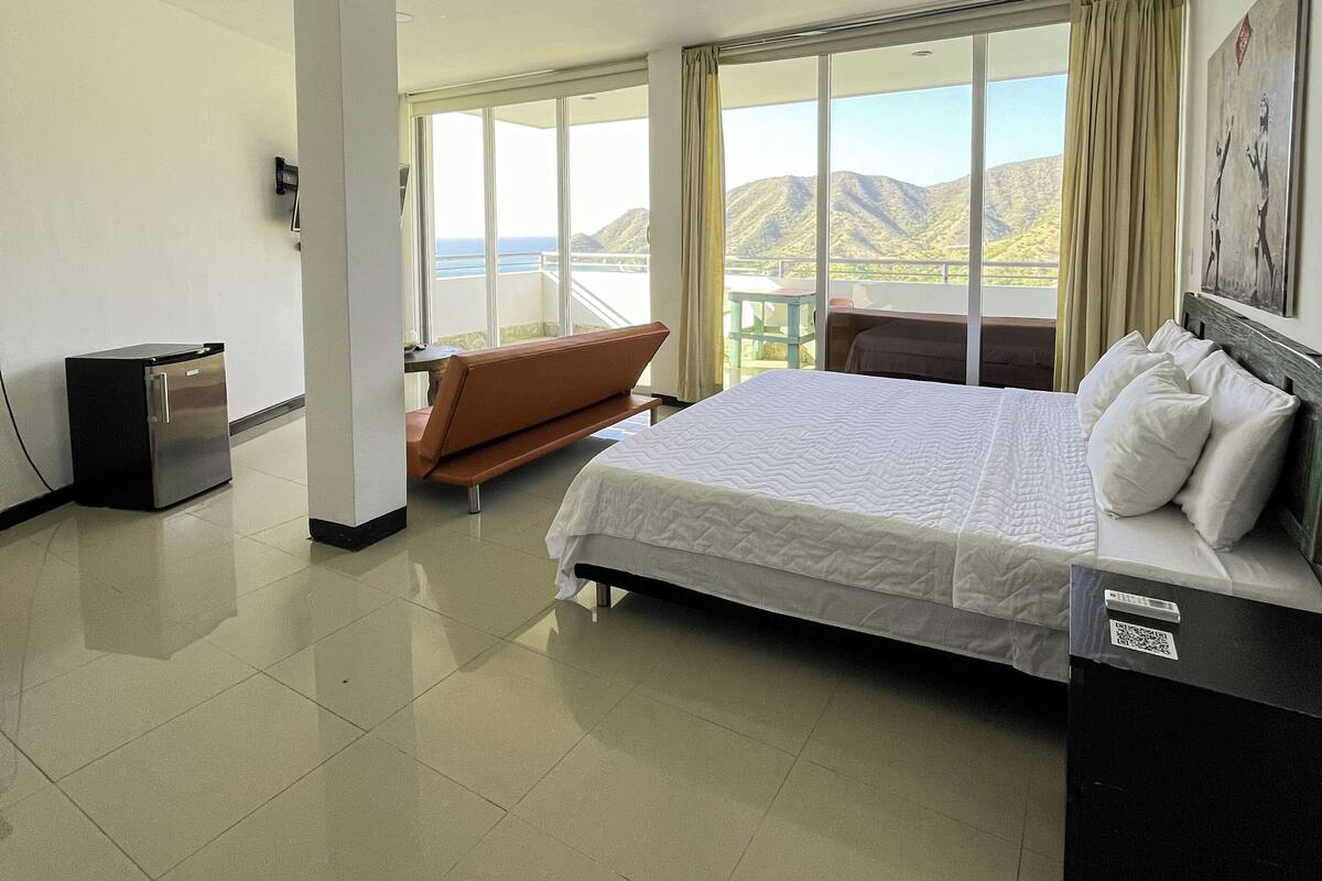 A hotel room with a bed, a small fridge, a seating area with a view, and a balcony.