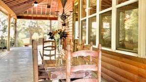 A wooden cabin with a covered porch, a table with chairs, and a window with a view of trees. - The CPL, 3BR Oxford Retreat by Velvet Ditch Villas (Batesville)