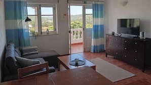 Et værelse med sofa, TV, bord og udsigt over naturen gennem en dør.