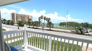 A balcony with white railings, two white lounge chairs with blue cushions, and a small table. In the background, there is a road, a stop sign, and a building with palm trees.