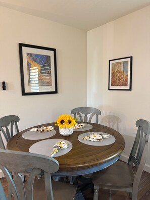 A dining area with a round table, six chairs, a vase with sunflowers, and two framed artworks on the walls.