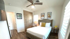 A bedroom with a wooden floor, a bed with white bedding, a ceiling fan, and a small table with a lamp.
