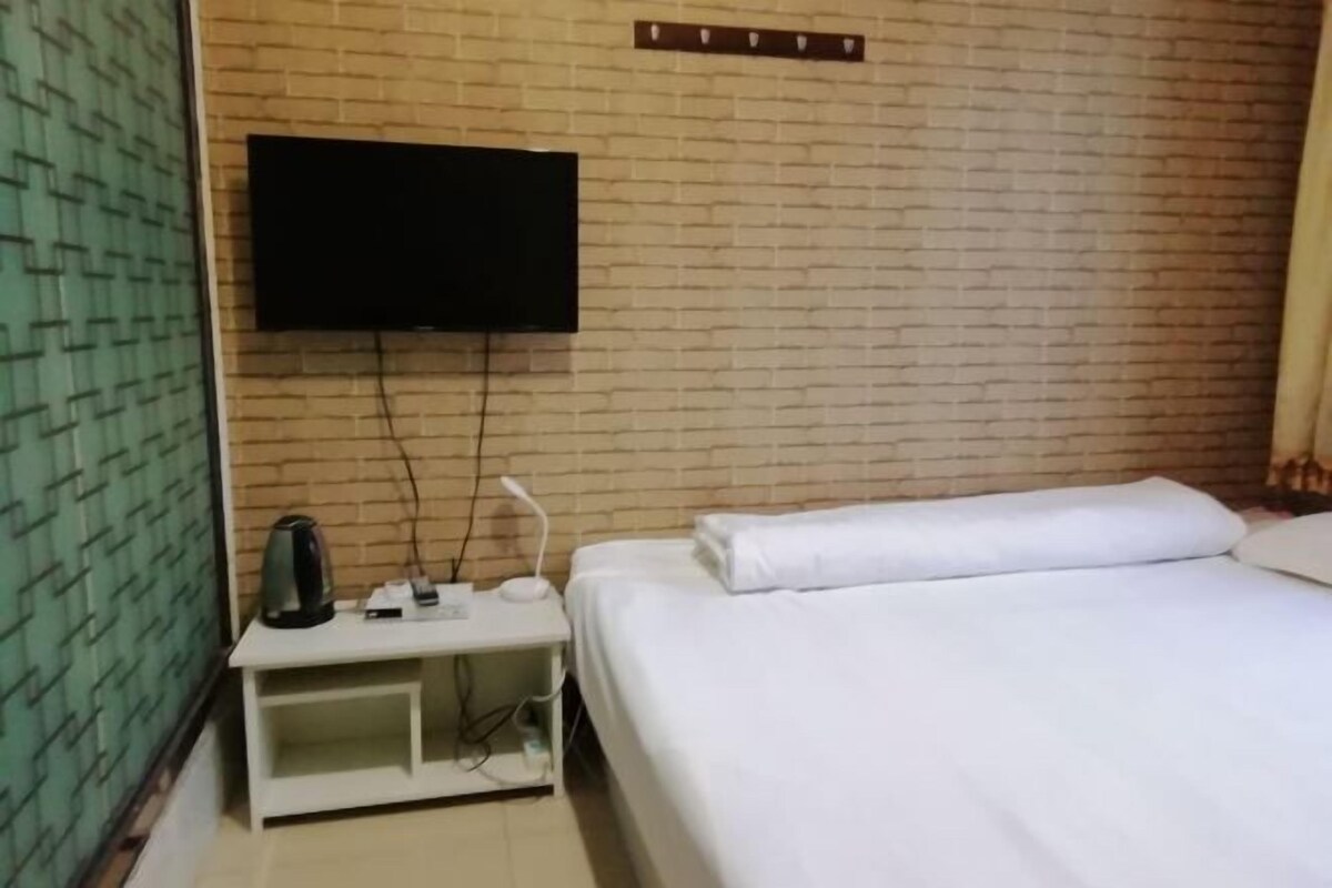 A hotel room with a brick-patterned wall, a flat-screen TV mounted above a white bedside table, and a neatly made bed with white linens.