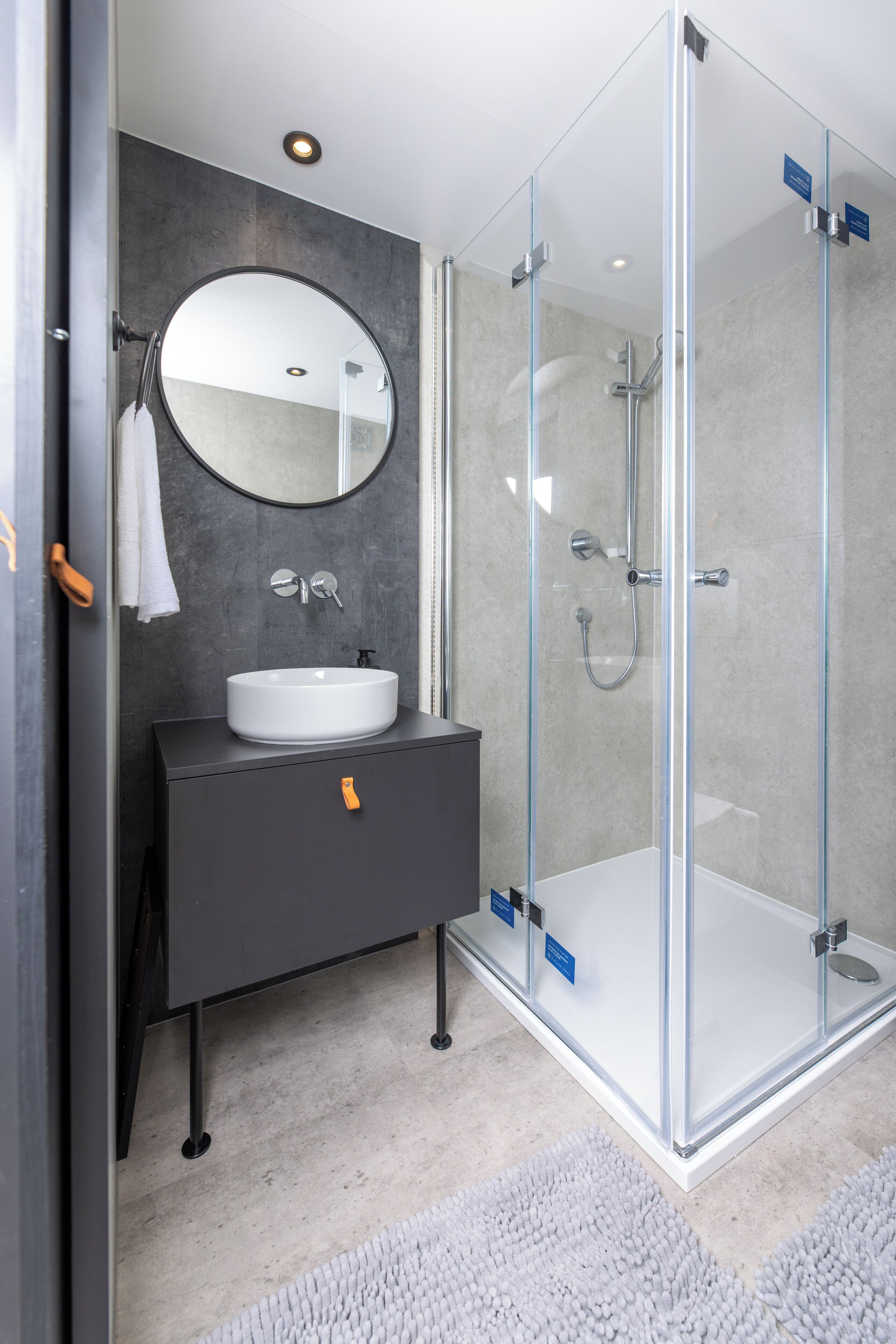 A modern bathroom with a glass-enclosed shower, a round mirror, and a dark vanity with a white sink.