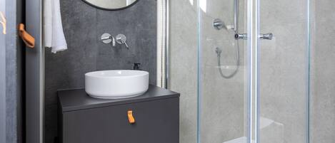 A modern bathroom with a glass-enclosed shower, a round mirror, and a dark vanity with a white sink.