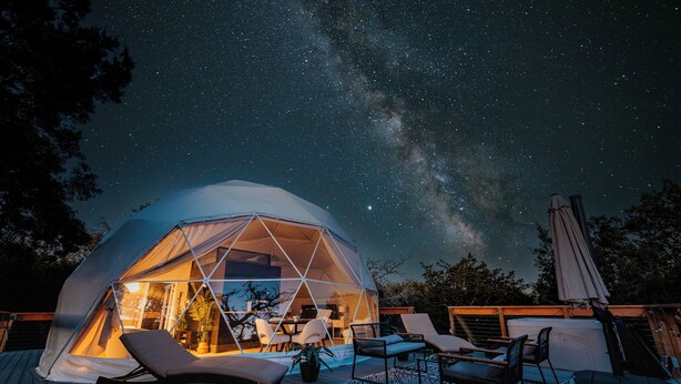 Une tente en forme de dôme géodésique, dotée d’un intérieur transparent et aménagée avec des chaises et une table, installée sur une terrasse en bois, sous un ciel étoilé.