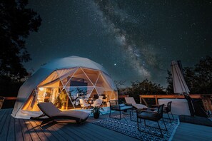 A geodesic dome tent with a clear interior, furnished with chairs and a table, set on a wooden deck under a starry night sky. - GAIA Geodome Fredericksburg (Fredericksburg)