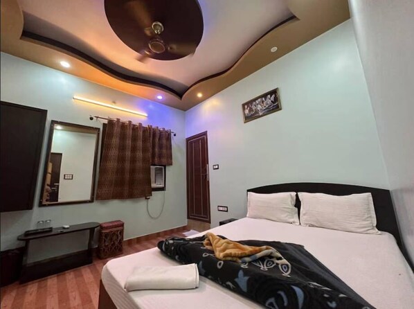A bedroom with a bed, a bedside table, a mirror, a wall-mounted air conditioner, and a ceiling fan. - Hotel Sai Palace (Varanasi)